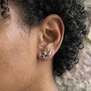 Puede incluir: Un pendiente de botón plateado en forma de llama es visible en la oreja. El pendiente tiene un acabado liso y pulido y un diseño de llama estilizado. La oreja está colocada sobre un fondo borroso.