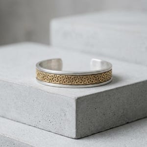 May include: A silver-toned cuff bracelet featuring a central band of gold-toned metal with an embossed repeating triquetra knot pattern. The bracelet has a smooth, polished finish on the edges and an open back design for easy adjustment.