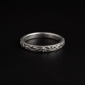 May include: A silver-toned metal ring featuring a continuous embossed pattern of delicate winding vines and small leaves. A single detailed bee motif is integrated into the botanical design, which wraps around the entire circumference of the band.