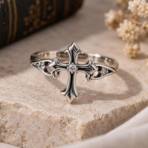 May include: A silver-toned metal ring featuring a central cross design with ornate, pointed edges. The cross is accented with a small, clear, round-cut gemstone at its centre, and two additional smaller clear gemstones are set into the band.