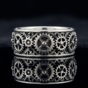 May include: A silver-toned ring featuring a continuous band design of interconnected mechanical gears and cogs. The metal surface has a dark, oxidized finish within the recessed areas to highlight the intricate details of the gear teeth and spokes.