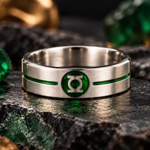 Op de afbeelding: Een zilverkleurige metalen ring met een geborstelde afwerking en een centrale horizontale groene groef. De band toont een rond embleem met een groene achtergrond en een zilverkleurig lantaarnsymbool in het midden van de ring.