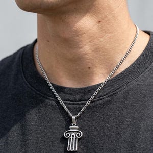 Op de afbeelding: Zilverkleurige ketting met een hanger in de vorm van een klassieke zuil. De hanger heeft ingewikkelde details, waaronder een kapiteel met scroll-achtige ontwerpen en verticale lijnen. De ketting is een curb-stijl.