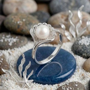 Pode incluir: Um anel de prata esterlina com uma grande pérola redonda em um design em forma de concha. O anel está sobre uma pedra azul, cercado por algas prateadas e uma estrela do mar. O fundo apresenta seixos e areia.