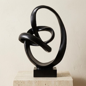 May include: A matte black sculpture featuring a continuous, fluid, knotted loop design. The object is mounted on a solid, rectangular black base, creating an abstract, interconnected form with smooth, rounded surfaces throughout the entire structure.