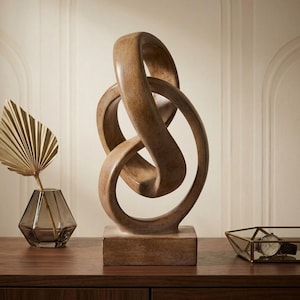 Puede incluir: Una escultura de madera marrón con un diseño abstracto de bucles continuos montada sobre una base rectangular. La superficie muestra una textura de veta de madera natural con bordes lisos y curvos que forman una forma compleja y anudada.