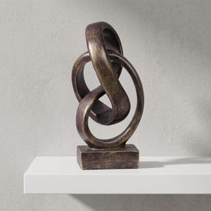 May include: Bronze-colored abstract sculpture with a looped design. The sculpture has a rectangular base and a textured finish. The artwork is displayed on a white shelf against a neutral gray background. The sculpture's dimensions are not visible.