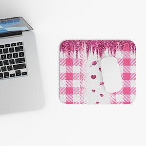 Puede incluir: Una alfombrilla de ratón a cuadros rosas y blancos con un ratón blanco. El borde superior tiene un diseño de purpurina rosa. Corazones y flechas rosas están dispersos sobre el fondo blanco. Un portátil está en el fondo.
