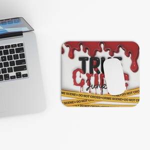 Puede incluir: Alfombrilla de ratón rectangular con el texto "True Crime Junkie" en rojo y negro, con un diseño de goteo de sangre. Un ratón blanco descansa sobre la almohadilla, y cinta amarilla de la escena del crimen bordea la parte inferior.
