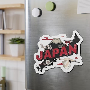 May include: A magnet featuring a map of Japan with Mount Fuji, cherry blossoms, and a crane. The word "JAPAN" is in red, with a distressed font. The magnet is on a silver refrigerator.