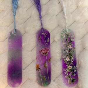 Op de afbeelding: Drie harsachtige bladwijzers met bloemmotieven en kwastjes. De bladwijzers hebben een verloop van paarse en groene tinten. Eén heeft een roze roos, een andere witte bloemen en de derde een aquareleffect. Elk heeft een kwastje in een andere kleur.