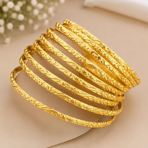 May include: A stack of eight gold-colored bangles, each featuring a textured, repeating pattern along the surface. The circular bracelets are arranged in a cascading formation, showcasing the metallic finish and the intricate, embossed detailing on every individual piece.