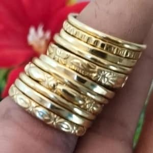 以下が含まれることがあります： 金色のリングのスタック。一部には、エッチングされた花柄や幾何学模様があります。指に重ねてあり、背景にはぼやけた赤い花があります。リングはデザインと質感が異なります。