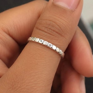 May include: A thin, polished silver-toned ring featuring a continuous band of small, rounded, square-shaped segments. The metal has a reflective, metallic finish, and the design consists of a repeating pattern of individual, slightly raised, square-like links that encircle the entire circumference of the band.