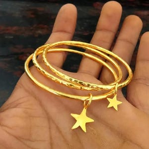 May include: Three gold-coloured bangles with a textured, hammered surface. Two of the bangles feature a small, flat, five-pointed star charm dangling from the circular band. The bracelets are thin, metallic, and have a polished, reflective finish.