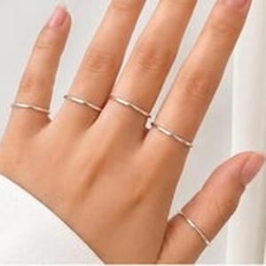 Pode incluir: Um conjunto de cinco anéis finos de metal em tom prateado polido usados nos dedos. Cada anel apresenta um design de banda simples, liso e esguio com uma subtil ponta em forma de V no centro, criando uma aparência uniforme e delicada na mão.