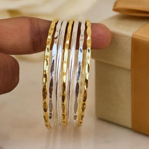 May include: A set of six thin bangles featuring alternating gold and silver tones. Each bangle has a distinct hammered, textured surface with small indentations along the outer edge, creating a reflective and metallic appearance for the jewelry.