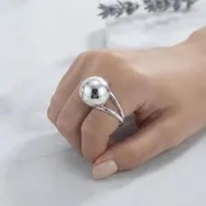 May include: A polished silver-tone ring featuring a large, smooth spherical bead centred on a split-shank band. The metallic surface of the sphere reflects light, creating a bright highlight, while the band divides into two distinct arms.