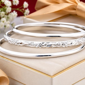 May include: A set of three silver bangles, including two plain polished bands and one central band featuring an intricate embossed floral pattern. The bracelets are arranged in a stack, showcasing a reflective metallic finish and rounded edges.