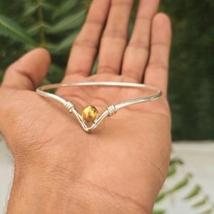 Puede incluir: Una pulsera rígida de tono plateado con un diseño central en forma de V y una cuenta esférica de tono dorado pulido. La banda metálica presenta pequeños detalles de alambre enrollado a ambos lados de la cuenta central, logrando un aspecto limpio.