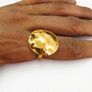 May include: A gold-toned ring featuring a large, circular, hammered metal face with an irregular, organic shape. The surface of the disc is highly reflective and smooth, catching light across its uneven, textured, and slightly concave design.