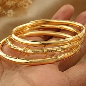 Könnte beinhalten: Ein Set aus drei goldfarbenen Armreifen. Zwei der Armreifen haben eine glatte, polierte Oberfläche, während der mittlere Armreif eine gehämmerte, strukturierte Oberfläche mit zwei kleinen, dekorativen goldfarbenen Ringen aufweist.