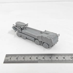 Op de afbeelding: Een grijs model van een militair voertuig met meerdere wielen en gedetailleerde componenten. Het model staat naast een liniaal met millimeter-metingen.