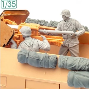 May include: A detailed model kit depicting a military scene. Two figures in uniform are positioned near a tan-colored vehicle, with a large gun barrel. The scene includes gray rolled-up bedrolls and the number "1/35".