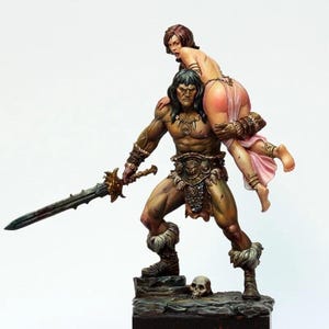 May include: A hand-painted miniature figurine depicting a muscular warrior with long dark hair, wearing a fur-trimmed loincloth and bracers, carrying a person over their shoulder. The warrior holds a long sword and stands on a rocky base with a skull.