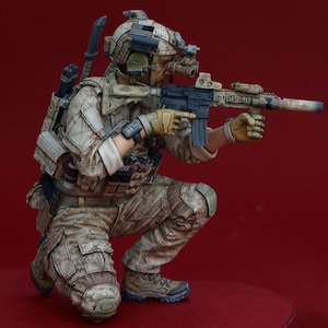 May include: A detailed figurine of a soldier in a combat pose, kneeling and aiming a rifle. The figure is dressed in desert camouflage, with tactical gear including a helmet, night vision goggles, and gloves. The rifle is black and tan. The background is a solid red.