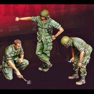 May include: Miniature figures of three soldiers in green uniforms and helmets. One is pointing, another kneeling, and the third bent over, possibly lighting a cigarette. The figures are set against a dark background.