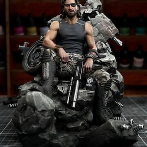 Könnte beinhalten: Detaillierte Figur eines Mannes mit Augenklappe, der ein graues Tanktop und eine Camouflage-Hose trägt und auf einem Motorrad-Thron sitzt. Er hält ein Gewehr. Die Figur ist von detaillierten, felsartigen Strukturen umgeben.