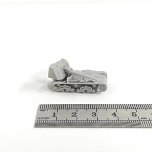 May include: A small, gray model of a tracked military vehicle with a large gun barrel and a sloped front. The model is positioned next to a ruler, showing millimeter measurements. The vehicle has detailed tracks and a textured surface.