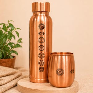 Puede incluir: Un juego de botella de agua y vaso de cobre. La botella y el vaso tienen un acabado brillante y presentan símbolos de chakra negros. La botella es alta y cilíndrica, mientras que el vaso es corto y redondeado.