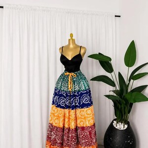 Peut inclure: Un haut noir à fines bretelles associé à une longue jupe maxi à étages, composée de bandes horizontales de tissu vert, bleu, jaune et rouge, chacune ornée de motifs blancs distincts et d'un cordon de serrage jaune à la taille.