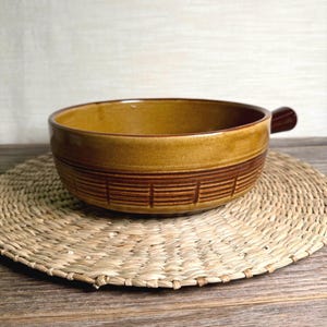 May include: A brown ceramic soup bowl with a handle. The bowl has a wide rim and a band of darker brown horizontal lines around the middle. It sits on a woven straw placemat on a wooden surface.