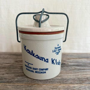 May include: A cream-colored ceramic crock with a wire bail handle and a matching lid featuring a red rubber seal. The front displays blue text reading Kaukauna Klub, It Spreads Like Butter, Copyright 1933, and Manufactured by Kaukauna Dairy Company.