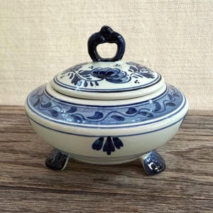 May include: A round ceramic lidded bowl featuring a cream-colored base with hand-painted dark blue floral and vine motifs. The lid has a decorative loop handle, and the bowl is supported by three small, dark blue curved feet at the base.