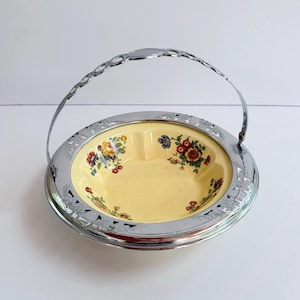 May include: A round, cream-colored ceramic bowl featuring a colorful floral pattern of yellow, red, and blue flowers. The bowl is set within a polished silver-tone metal frame that includes a decorative, arched handle with a cutout design.