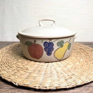 May include: A beige ceramic casserole dish with a lid and two handles. The dish is decorated with painted illustrations of a red apple, blue grapes, and a yellow pear. The dish is sitting on a woven placemat.
