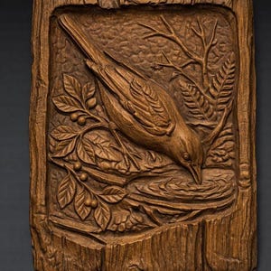 May include: A detailed, brown wooden carving of a bird drinking water from a small pool. The bird is surrounded by leaves, berries, and branches, all carved into a rectangular wooden frame. The wood grain is visible.