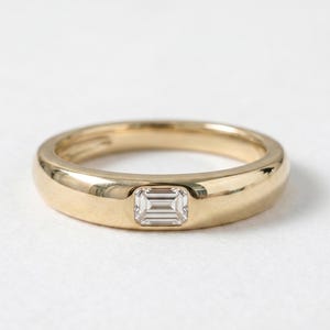 Può includere: Un anello a fascia in oro lucido con una singola pietra preziosa trasparente taglio smeraldo, incastonata orizzontalmente a filo della superficie metallica. L'anello presenta un profilo liscio e arrotondato con una finitura lucida.