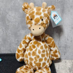 May include: A plush giraffe toy with a brown and white spotted pattern. The stuffed animal has a tan snout, small ears, and a tag attached. The giraffe is sitting upright on a dark surface.