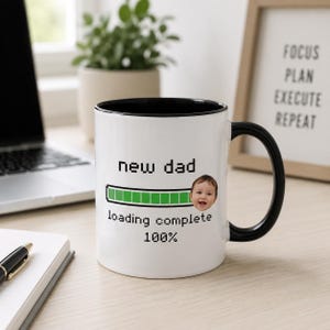 Peut inclure: Un mug en céramique blanche avec une anse et un bord intérieur noirs. L'avant présente un texte noir pixélisé indiquant new dad, une barre de chargement verte, une petite photo du visage d'un bébé et le texte loading complete 100%.