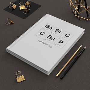 May include: A white notebook with the phrase "just basic crap" and a periodic table-style design. Gold and black pencils and binder clips are arranged around the notebook on a dark gray surface.
