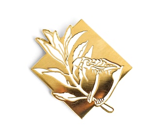 Può includere: Uno spillo in metallo color oro con un contorno bianco di un fiore e foglie. Lo spillo ha la forma di un diamante.
