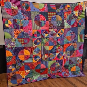 Könnte beinhalten: Ein farbenfrohes Patchwork-Quilt mit einem lebendigen Muster. Es zeigt eine Vielzahl von Stoffen in Rosa, Blau, Orange, Grün und Lila. Das Design umfasst Quadrate und Halbkreise, die ein lebendiges Muster bilden. Der Quilt wird vor einem neutralen Hintergrund präsentiert.