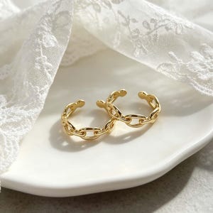 May include: Two gold-colored chain link rings are displayed on a white plate, set against a backdrop of delicate white lace. The rings have an open design, allowing for adjustable sizing. The warm lighting enhances the gold's shine.