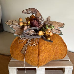 Könnte beinhalten: Eine handgefertigte orangefarbene Filzkürbis-Dekoration mit einem gebogenen braunen Stiel, Stoffblättern mit rissiger Textur und kleinen gelben Beeren. Ein kleines Holzschild mit der Aufschrift "fatto a mano" ist mit einer Schnur befestigt.