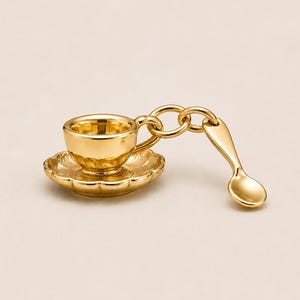 May include: A gold-coloured charm depicting a miniature teacup, saucer, and spoon. The teacup rests on a scalloped saucer, and the spoon is suspended from a chain attached to the teacup. The charm is made of a shiny, reflective material.